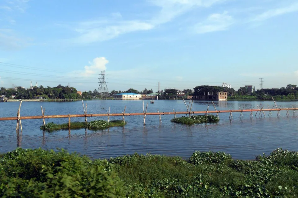 Monsoon floodwater spreading across low land in Narsingdi with power lines and buildings beyond