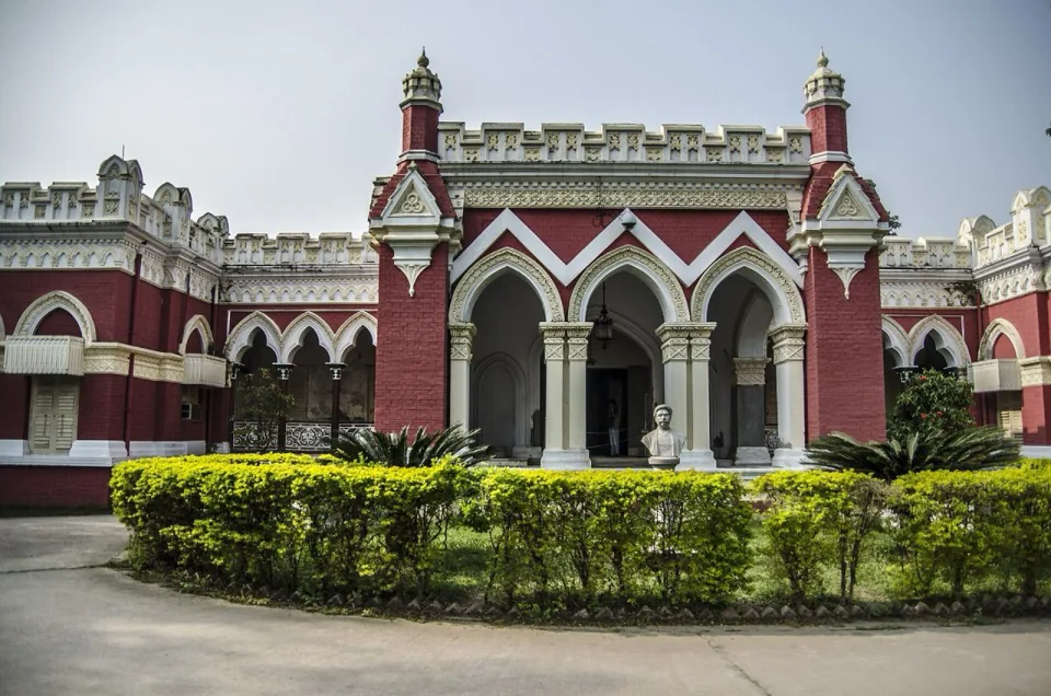 Red and white arched facade of the Uttara Ganabhaban palace behind a hedge